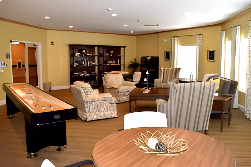 A senior living common room with upholstered armchairs, a shuffleboard table, a round table in the foreground, and a display cabinet along the back wall.