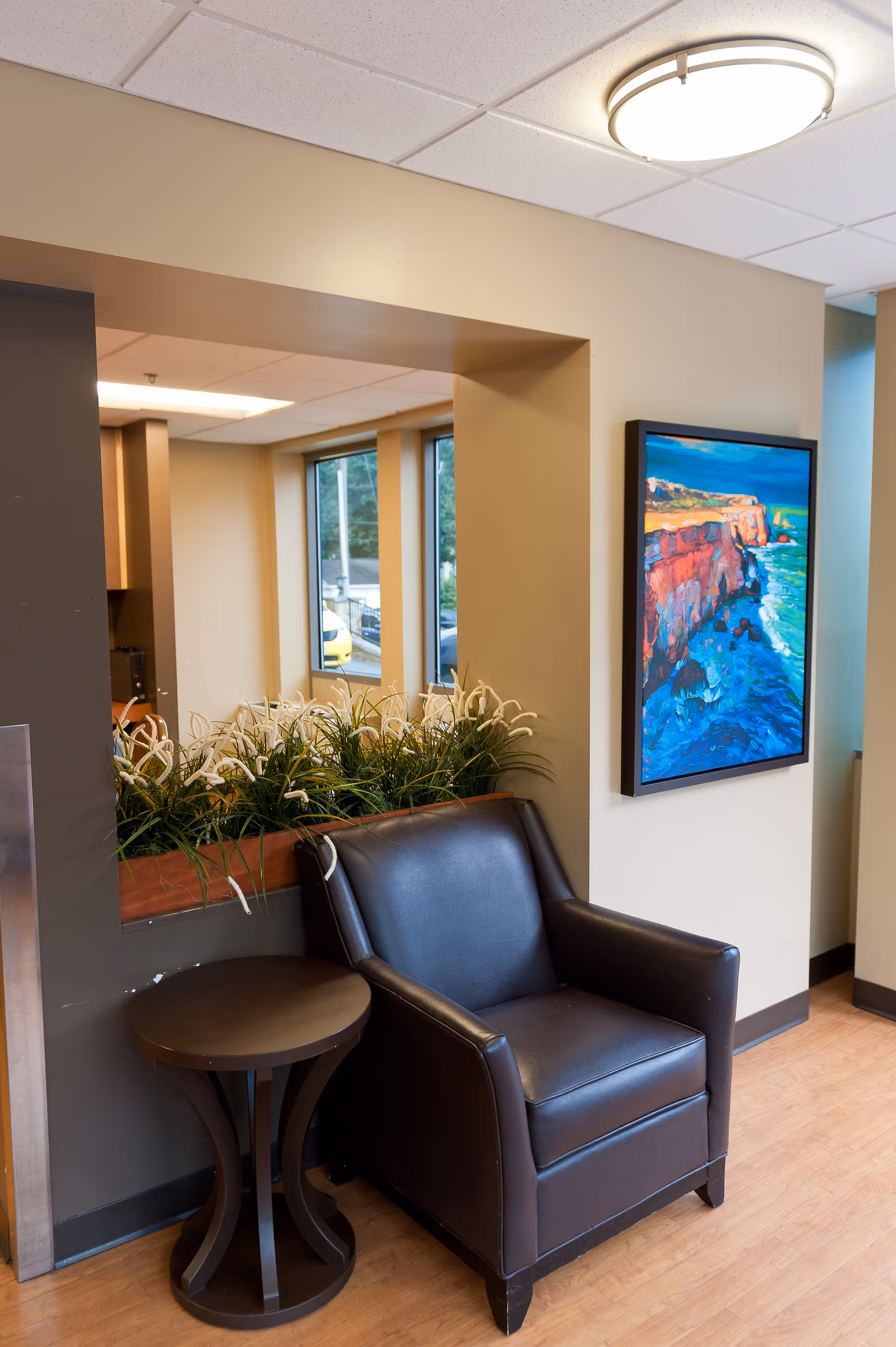 A leather armchair and side table next to a planter and colorful seaside painting in a facility waiting area.