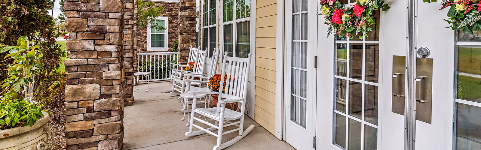 A covered outdoor porch area with white rocking chairs lined up along the wall next to large windows and glass doors. The porch features stone pillars and potted plants with flowers, creating a welcoming and relaxing atmosphere.