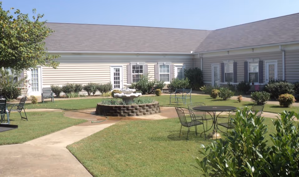 Outdoor courtyard area of a senior living facility with a central stone fountain surrounded by green grass, metal tables and chairs, bushes, and a beige building with multiple windows and doors in the background under a clear blue sky.