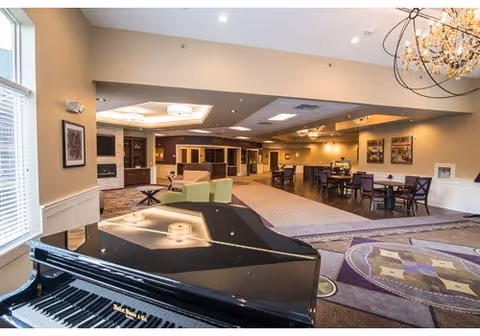 Spacious and well-lit common area in a senior living facility featuring a black grand piano in the foreground, several seating areas with chairs and tables, decorative lighting fixtures, and large windows allowing natural light to enter.