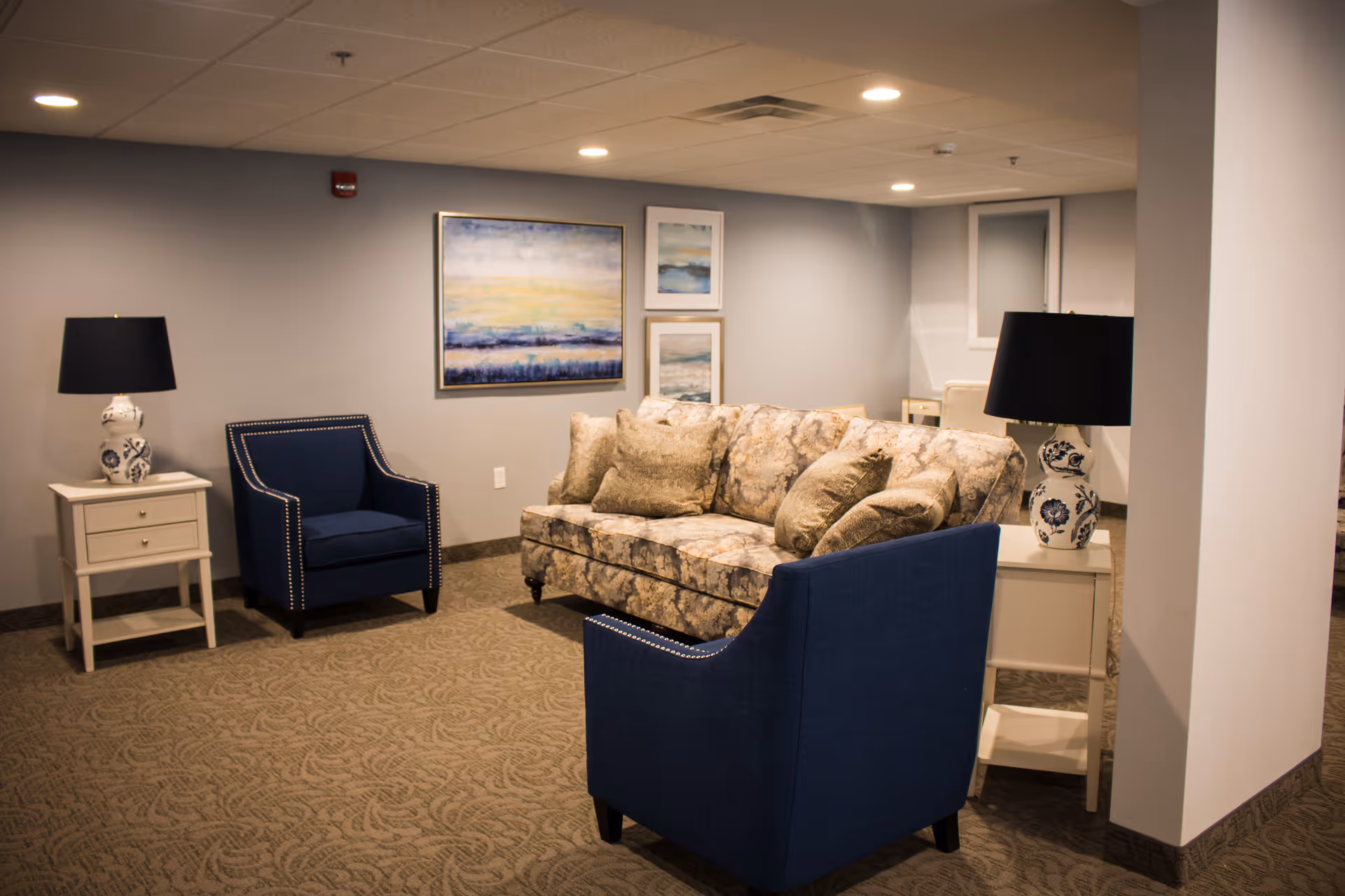 A cozy living room area with a floral-patterned sofa and two blue armchairs with nailhead trim. There are two white side tables, each with a decorative lamp featuring a white base with blue floral designs and black lampshades. The walls are light gray and decorated with three framed abstract paintings. The floor is carpeted with a patterned design, and the ceiling has recessed lighting.