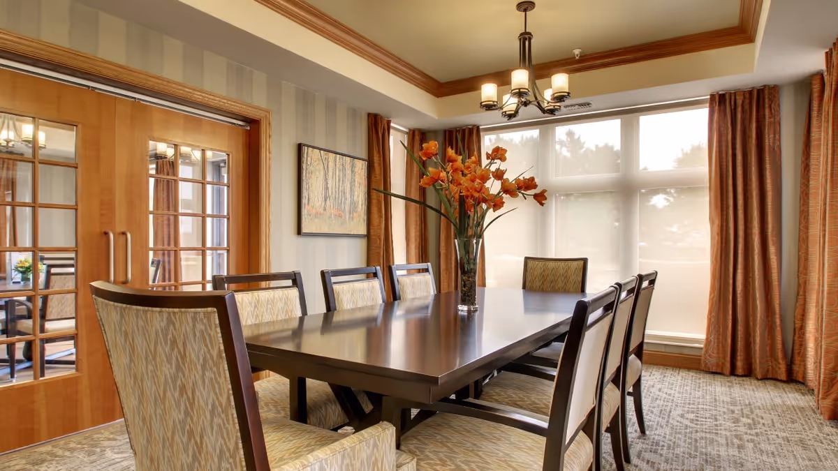 A well-lit dining room with a large dark wooden table surrounded by eight upholstered chairs. A vase with orange flowers is placed in the center of the table. The room features large windows with sheer white blinds and orange curtains, a chandelier hanging from the ceiling, and wooden framed glass double doors on one side. A framed painting is visible on the striped wallpapered wall.