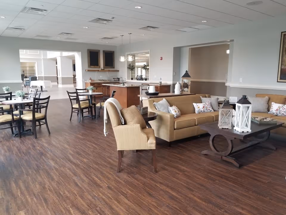 A spacious and well-lit common area in a senior living facility featuring a seating area with beige sofas and armchairs adorned with decorative pillows. A dark wooden coffee table with white lanterns sits in front of the sofas. To the left, there are several small round tables with chairs, and in the background, a kitchen island with pendant lights hangs above. The room has wood flooring and light-colored walls with large windows allowing natural light.