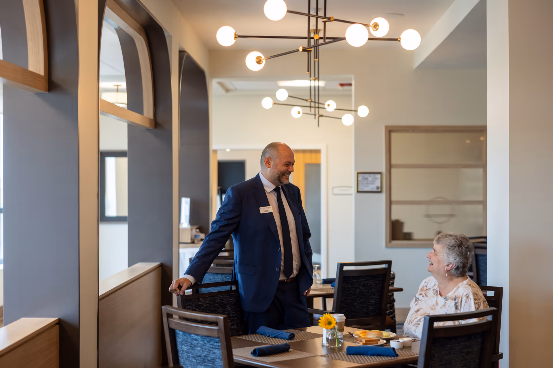 A man in a blue suit is standing and smiling while talking to an elderly woman seated at a dining table in a well-lit room with modern light fixtures and large windows.