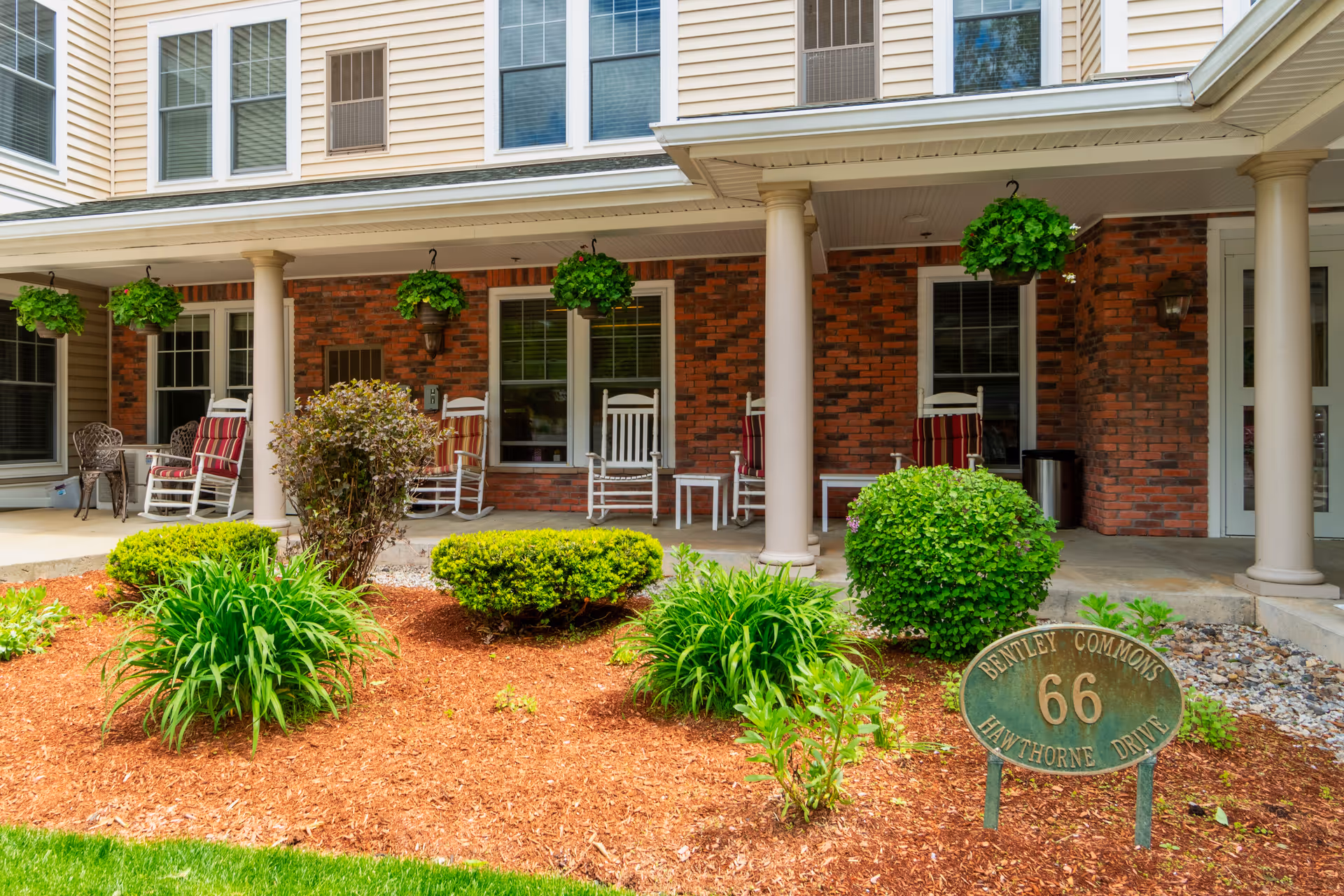 Front porch area of Bentley Commons at Bedford featuring a covered patio with white rocking chairs and hanging green plants. The building has a brick facade with beige siding above and several windows. In front of the porch is a landscaped garden with bushes, green plants, and a sign that reads 'Bentley Commons 66 Hawthorne Drive'.