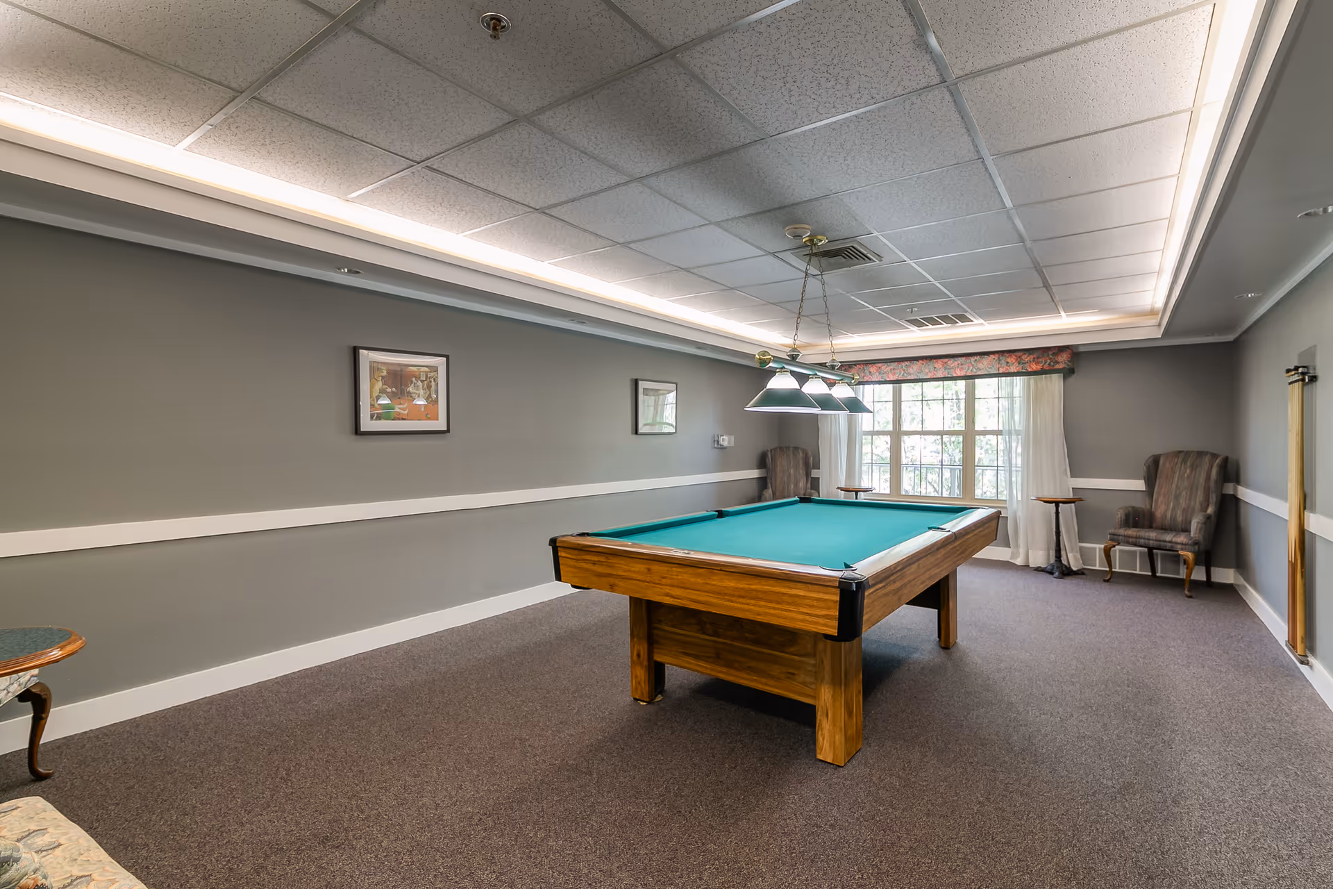 Interior game room with a pool table under hanging lights, upholstered chairs, and a large window.