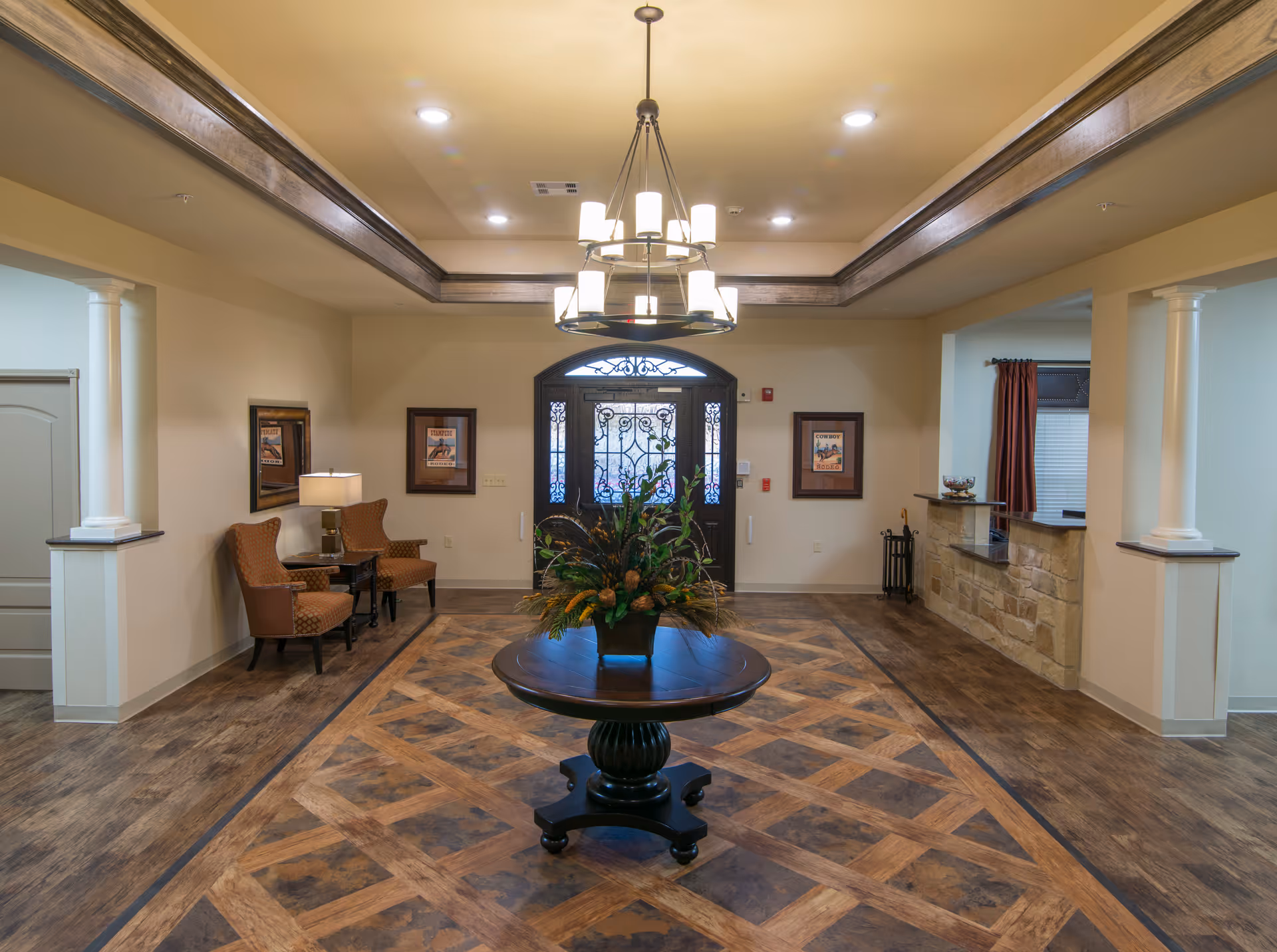 A spacious and well-lit lobby area with a decorative round wooden table in the center holding a floral arrangement. The floor has a patterned wood design, and the ceiling features a tray design with recessed lighting and a chandelier. There are two brown chairs with a small table and lamp on the left side, framed pictures on the walls, and a stone reception desk on the right. The entrance door is ornate with glass and wrought iron detailing.