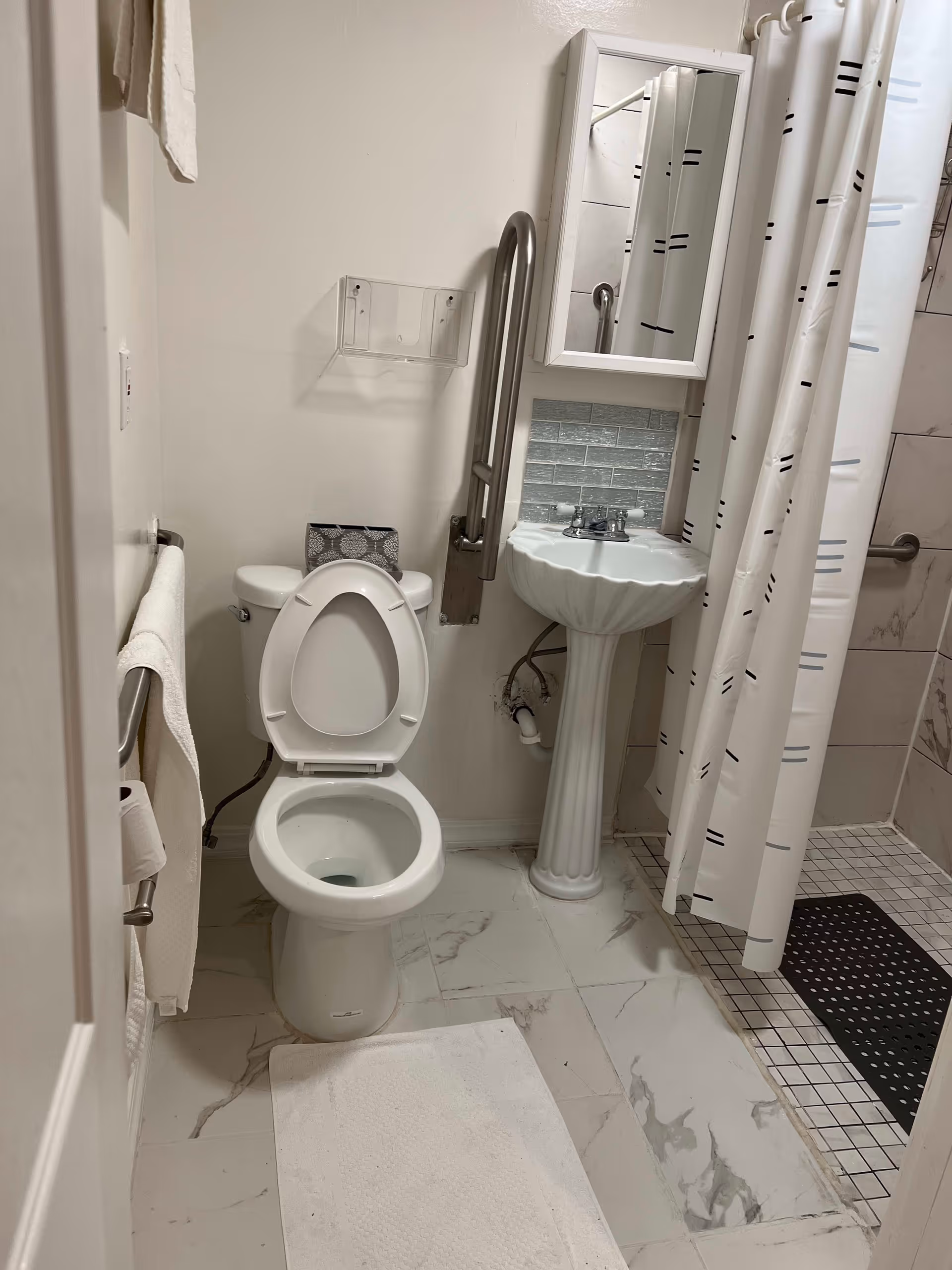 A clean bathroom with a white toilet with the lid open, a pedestal sink with a mirror above it, a shower area with a white curtain featuring black and blue lines, and grab bars installed near the toilet and in the shower for accessibility. The floor is tiled with a white and gray marble pattern, and a white bath mat is placed in front of the toilet.