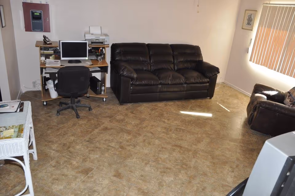 A living room with a black leather sofa and matching armchair, a computer desk with a monitor, keyboard, printer, and office chair, beige tiled floor, and a window with vertical blinds letting in sunlight.