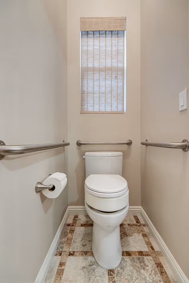 A small bathroom with a white toilet centered against the back wall. There are stainless steel grab bars on both side walls and behind the toilet. A toilet paper holder with a roll of toilet paper is mounted on the left wall. Above the toilet is a narrow window with a beige pleated shade partially covering it. The floor has a patterned tile design with beige and brown tones.