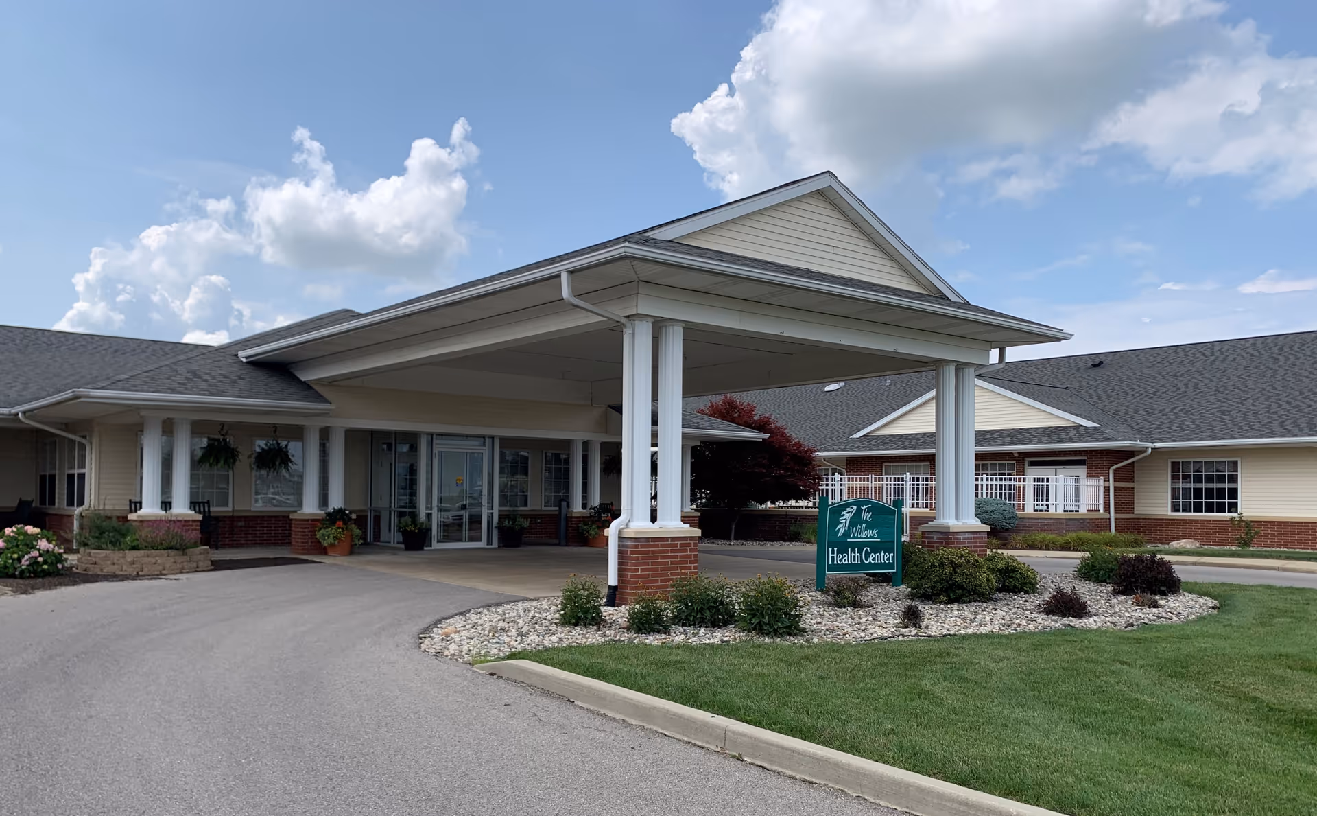 Exterior view of The Willows at Bellevue Health Center showing the entrance with a covered drop-off area supported by white columns. The building has a combination of brick and light-colored siding with a well-maintained lawn and landscaping around the driveway.