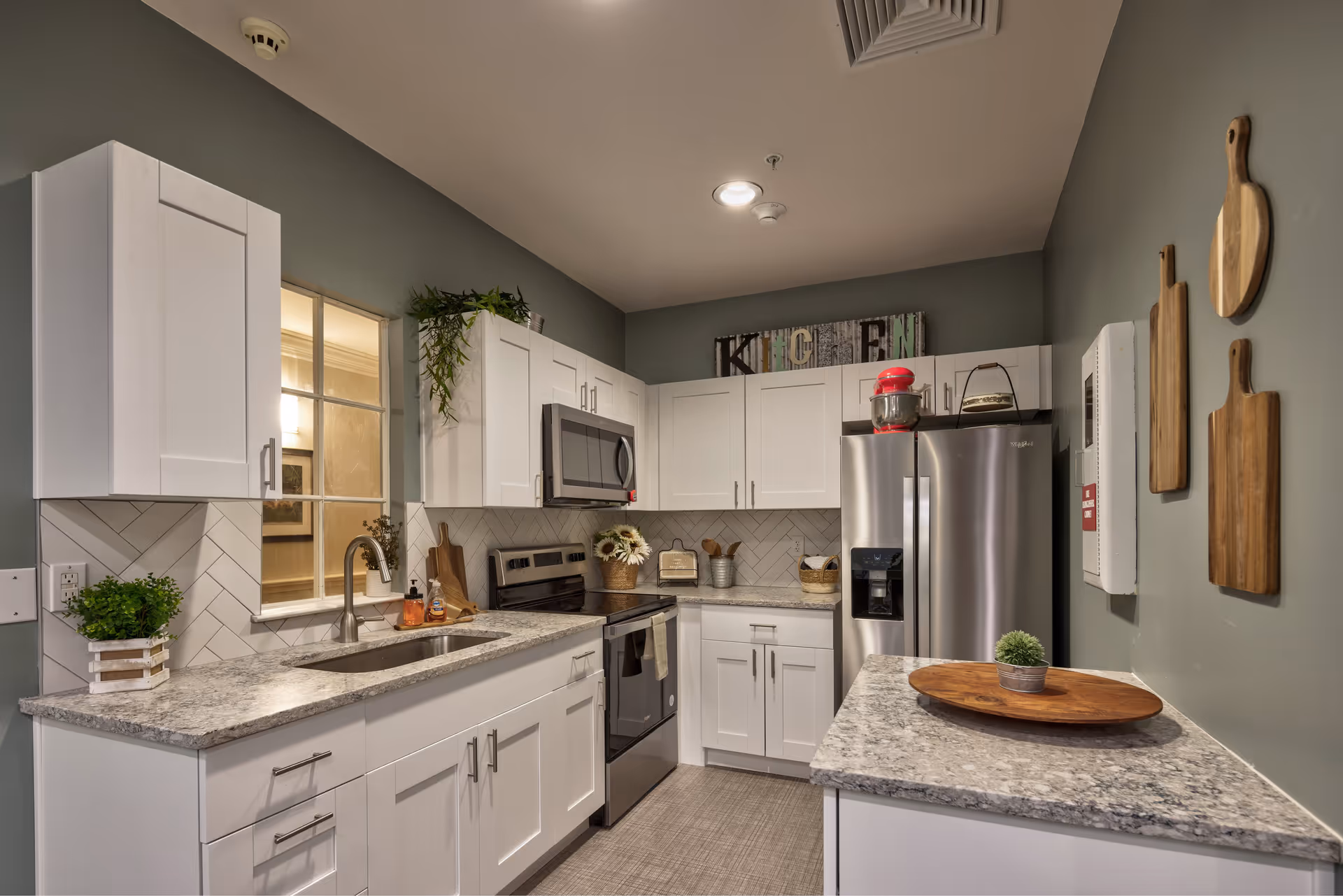 Modern kitchen with white cabinets, granite countertops, stainless steel appliances including a refrigerator, stove, and microwave. Decorative wooden cutting boards hang on the wall, and a small plant sits on a wooden tray on the counter. A sign spelling 'KITCHEN' is displayed above the cabinets.
