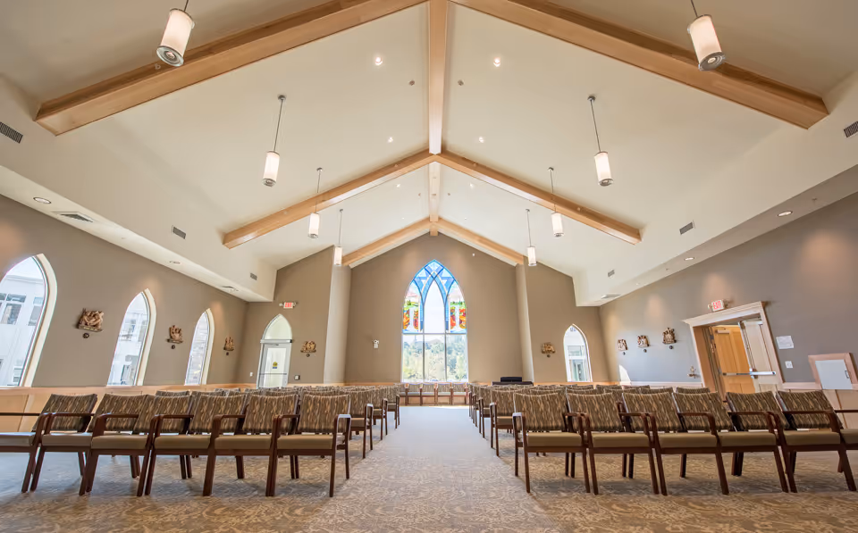 Interior view of a chapel or large meeting room with high vaulted ceilings and wooden beams. Rows of cushioned chairs are arranged facing a large stained glass window at the far end. The room has arched windows on the side walls and pendant lights hanging from the ceiling.