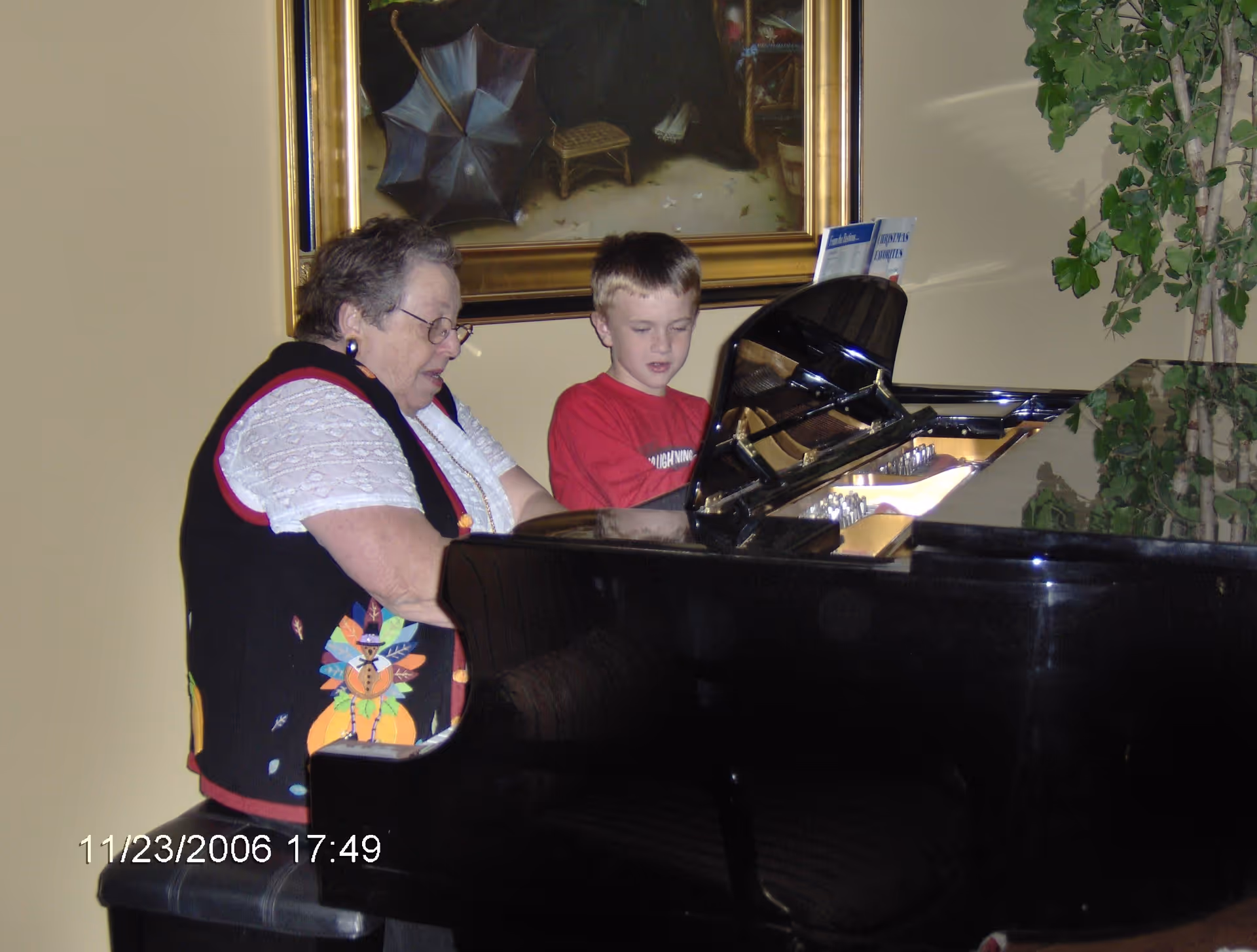 An elderly woman and a young boy sitting together at a black grand piano, with the woman playing and the boy watching. There is a large framed painting on the wall behind them and a tall green plant to the right.