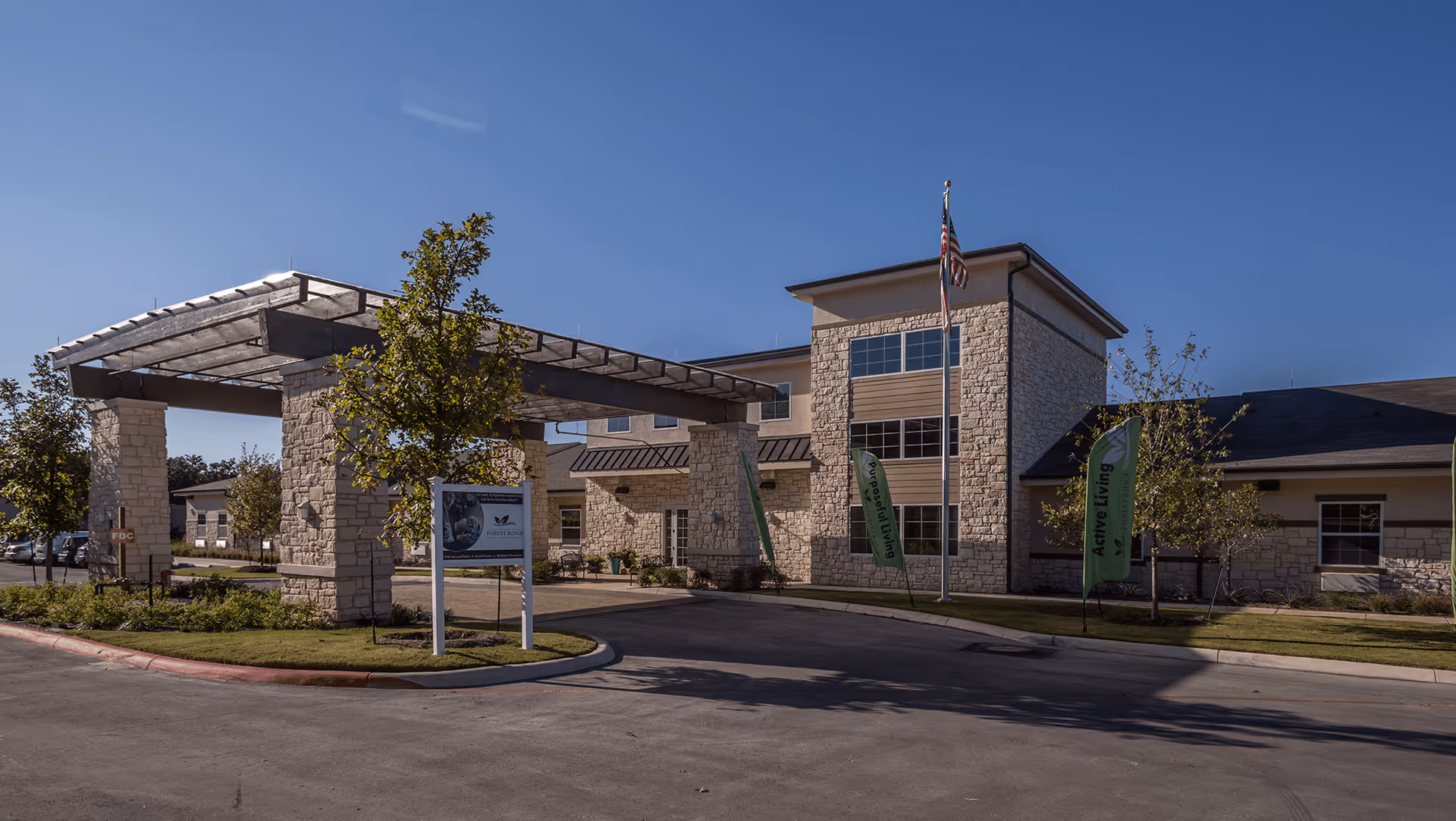 Exterior view of The Legacy at Forest Ridge senior living facility showing a stone building with a covered entrance, an American flag on a flagpole, green banners that say 'Active Living', trees, and a clear blue sky.