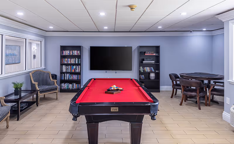 A recreational room with a red felt pool table in the center, two black bookshelves filled with books on the back wall, a large flat-screen TV mounted between the bookshelves, a round table with four chairs to the right, and two striped armchairs with a small table and plant to the left.
