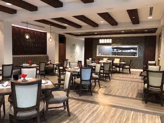 A spacious dining room with multiple tables and chairs arranged neatly. The room features wooden ceiling beams, pendant lighting, and a decorative wall with a built-in display. The floor has a wood-like finish, and the overall ambiance is warm and inviting.