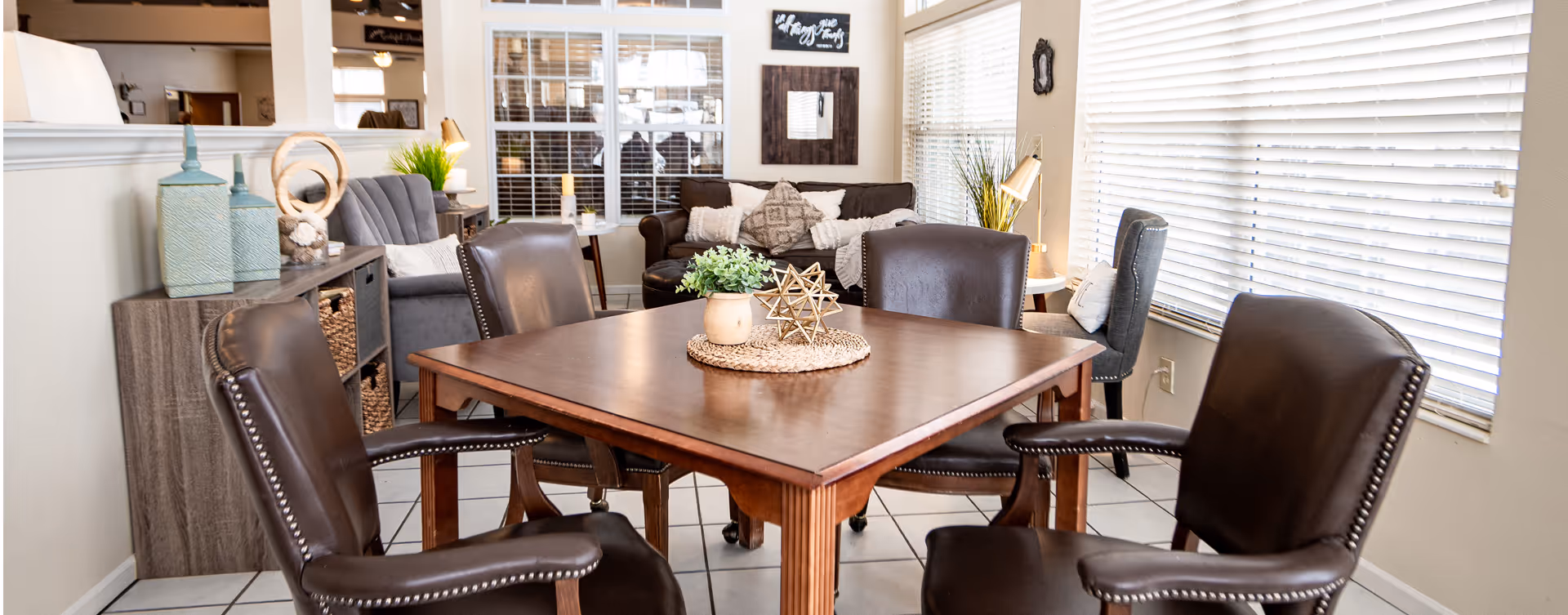 A cozy interior room with a wooden square table surrounded by four brown leather chairs. In the background, there is a gray sofa with decorative pillows, a gray armchair, and a side table with a lamp and plant. The room has large windows with white blinds allowing natural light to fill the space. Decorative items and plants are placed on a wooden cabinet and tables, creating a warm and inviting atmosphere.