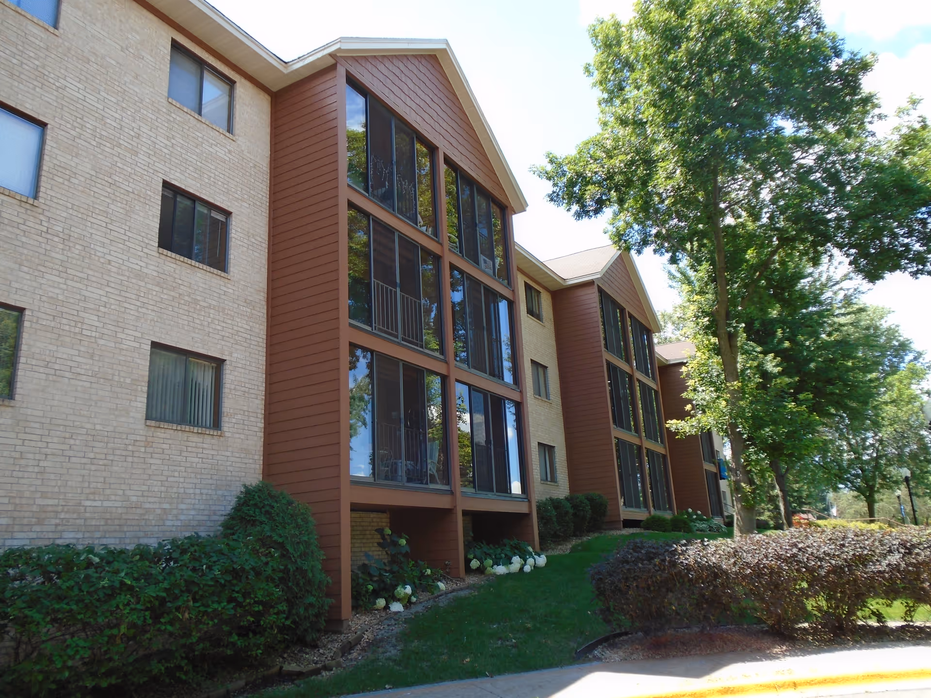 Exterior view of a multi-story senior living facility building with large windows and brown siding sections. The building is surrounded by green bushes, trees, and a well-maintained lawn under a clear sky.