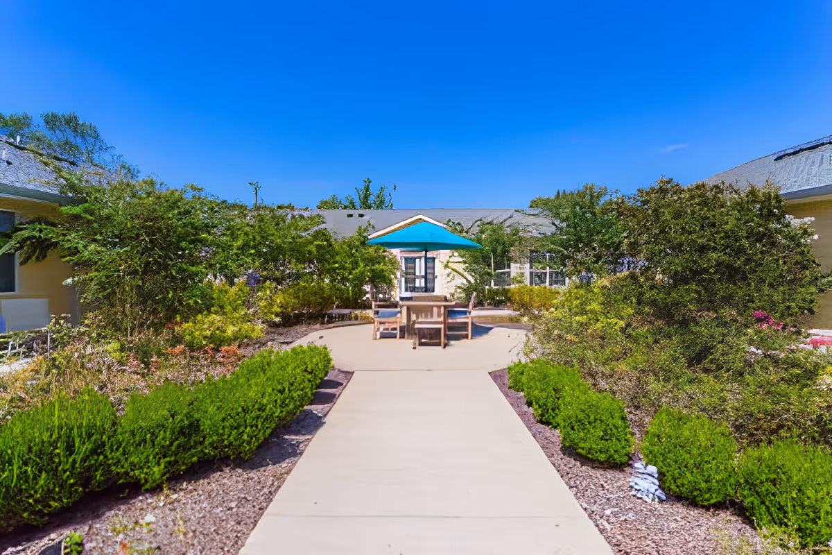 Outdoor courtyard area at Charter Senior Living of Hermitage with a concrete walkway leading to a seating area featuring a table and chairs under a blue umbrella, surrounded by green bushes and plants under a clear blue sky.