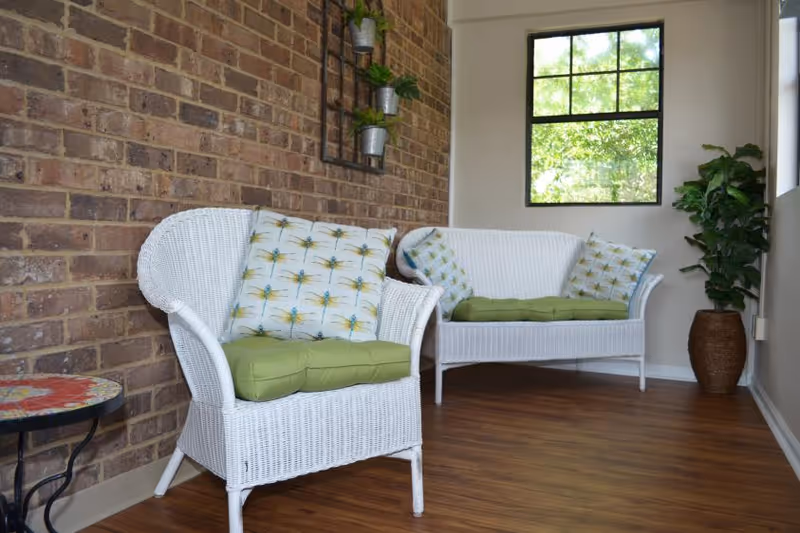 A cozy sitting area with white wicker furniture including a chair and a loveseat, both with green cushions and patterned pillows. The space features a brick wall with small potted plants mounted on a black metal frame, a window showing greenery outside, a wooden floor, and a tall potted plant in the corner.