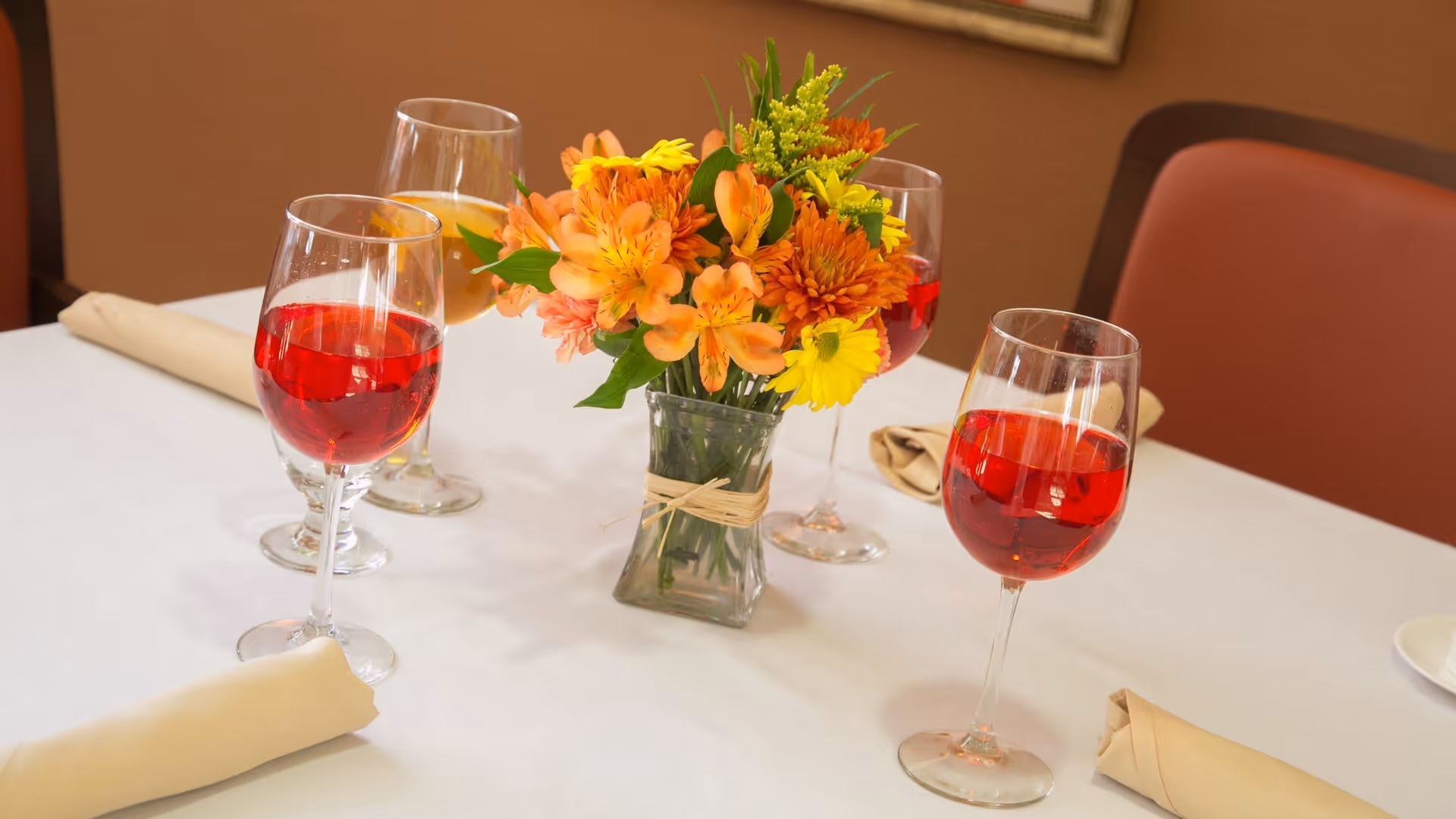 A dining table set with four glasses of red and white beverages, beige cloth napkins, and a small vase with a colorful bouquet of orange, yellow, and green flowers in the center.
