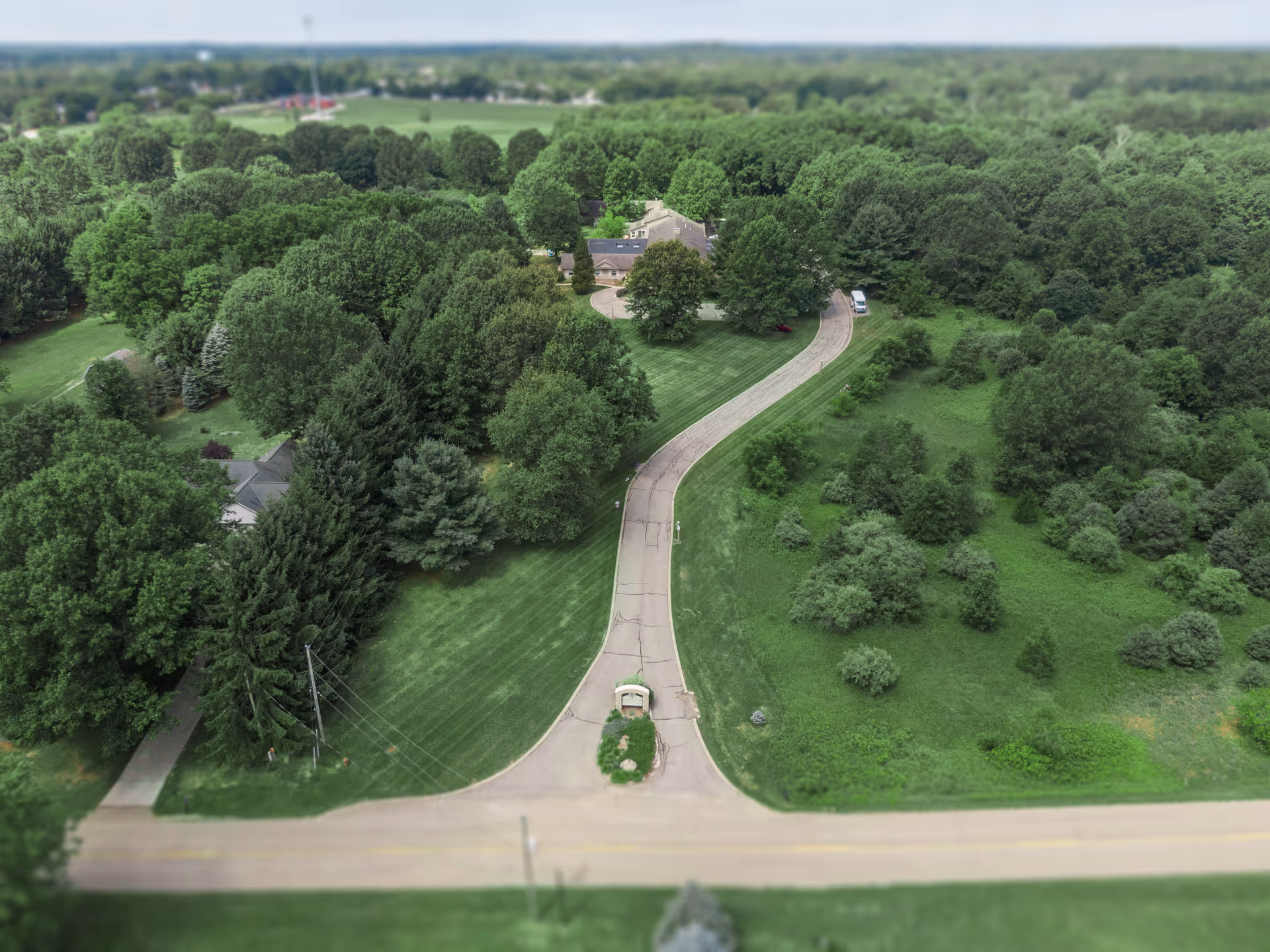 Aerial view of a winding paved driveway flanked by lawns and dense trees leading to a house set within a wooded property.