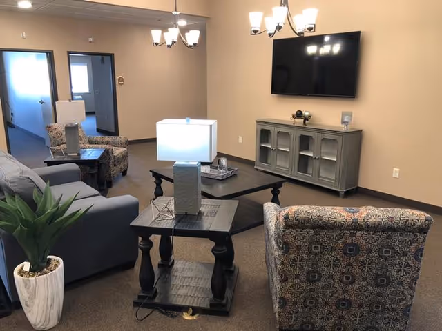 Community living room with upholstered chairs, sofas, side tables, a potted plant, and a wall-mounted TV.