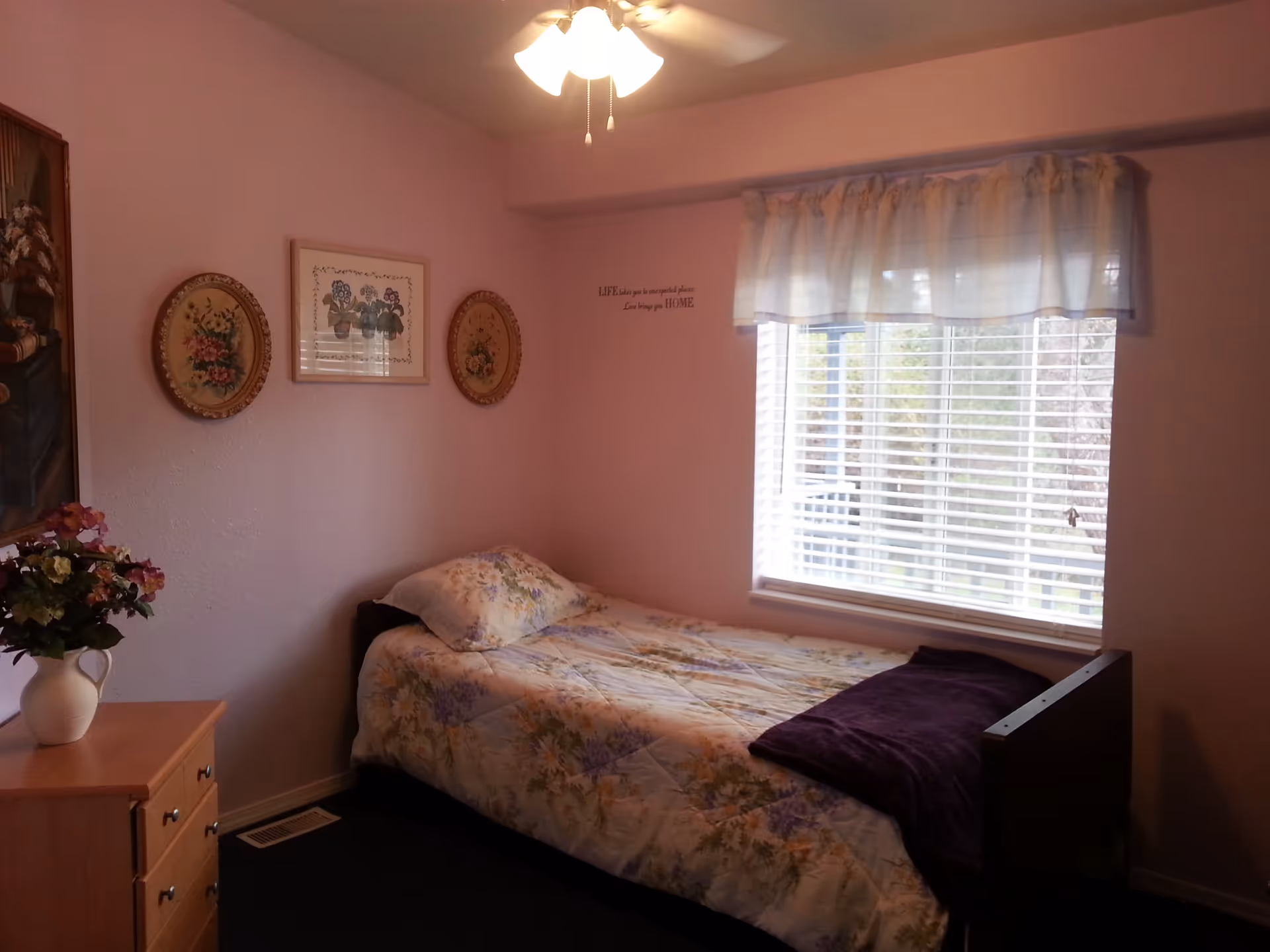 A small bedroom with a single bed covered in floral bedding and a purple blanket. There is a wooden dresser with a vase of flowers on it, three framed floral artworks on the wall, and a window with blinds and a sheer curtain letting in natural light. A ceiling fan with lights is visible above.