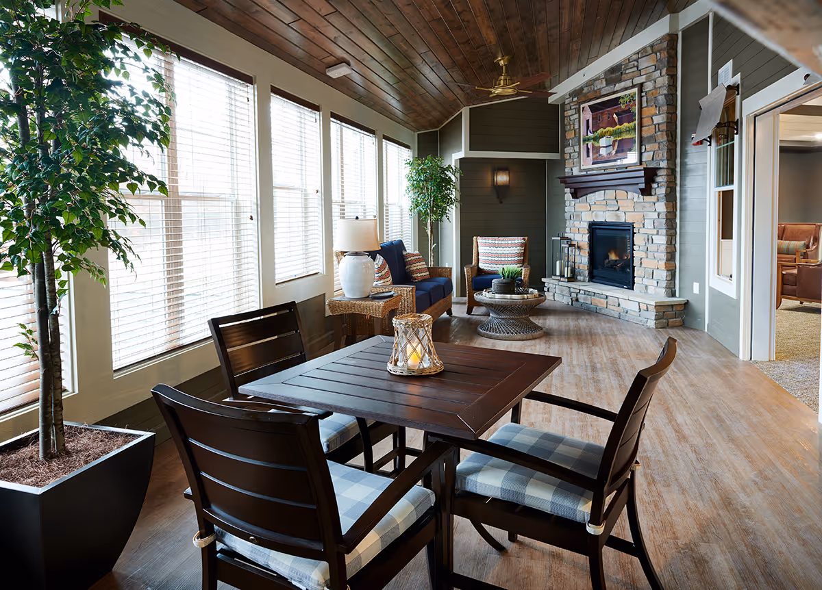A cozy indoor sitting area with a wooden table and four cushioned chairs in the foreground. Large windows with white blinds line the left wall, allowing natural light to fill the space. There are two potted plants, a table lamp on a side table, and a stone fireplace with a painting above it on the far wall. The ceiling is wooden with a ceiling fan, and an adjacent room with additional seating is visible through an open doorway.