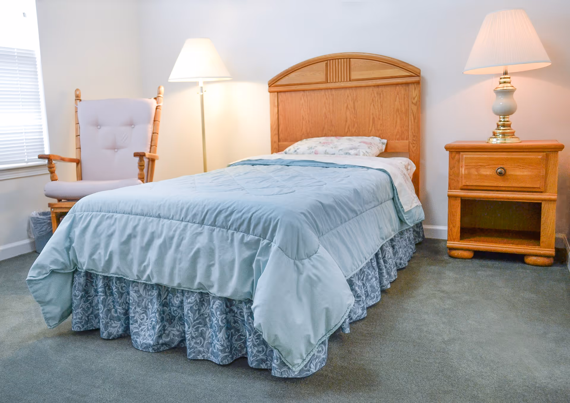 A neatly made single bed with a light blue comforter and floral pillow in a room. Next to the bed is a wooden nightstand with a lamp on it. To the left of the bed is a cushioned wooden rocking chair and a floor lamp. The room has light-colored walls and a window with blinds partially visible.