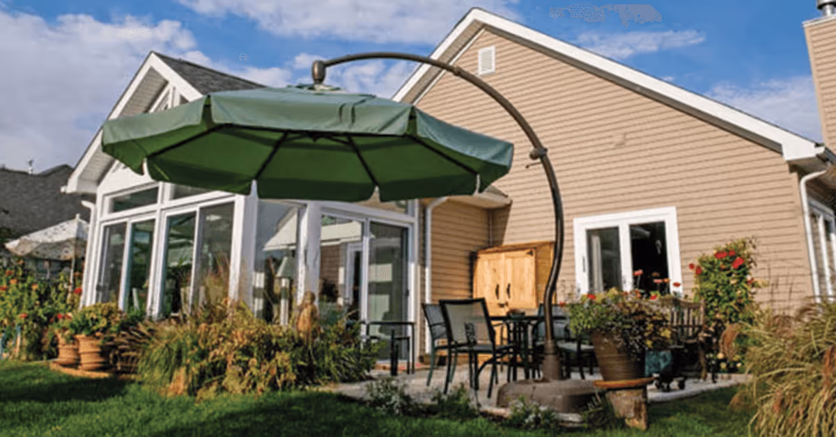 Outdoor patio area at Otterbein Granville SeniorLife Community with a large green umbrella, several chairs, a table, and surrounding plants and flowers next to a beige building with white trim under a partly cloudy sky.