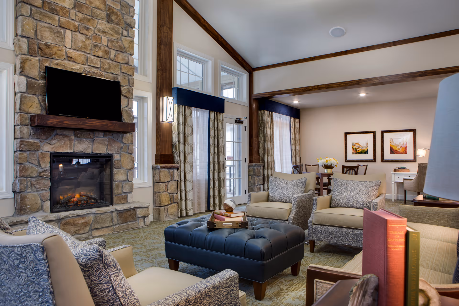 A cozy living room area with a stone fireplace and a mounted flat-screen TV above it. The room features beige and patterned armchairs arranged around a tufted dark blue ottoman with books on top. Large windows with patterned curtains allow natural light to enter. In the background, there is a dining table with chairs and framed artwork on the wall. The room has wooden beams and trim, creating a warm and inviting atmosphere.