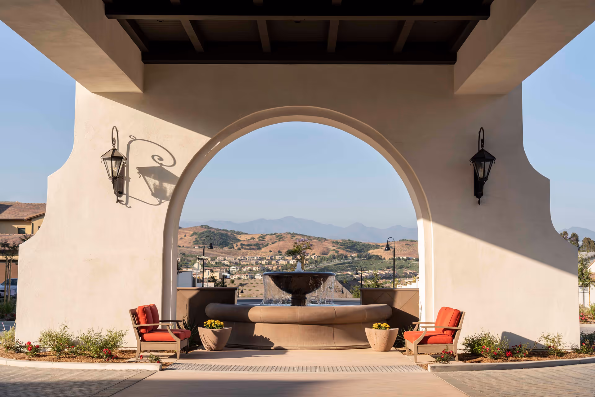 Outdoor seating area with two red cushioned chairs facing a large stone fountain under a covered archway, with hills and houses visible in the background under a clear sky.