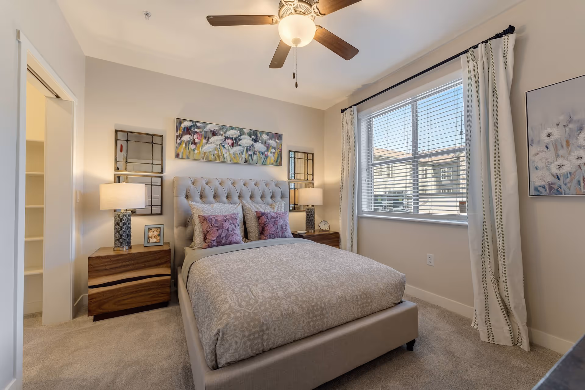 A cozy bedroom with a beige upholstered bed featuring patterned bedding and two purple pillows. There are two wooden nightstands on either side of the bed, each with a lamp. Above the bed is a floral painting and two decorative mirrors. A large window with white curtains lets in natural light, and a ceiling fan with a light fixture is mounted on the ceiling.