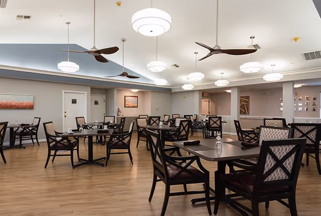 A spacious dining room with multiple dark wood tables and chairs arranged neatly on a wooden floor. The ceiling features modern round light fixtures and ceiling fans. The walls are light-colored with framed artwork and doors visible in the background.