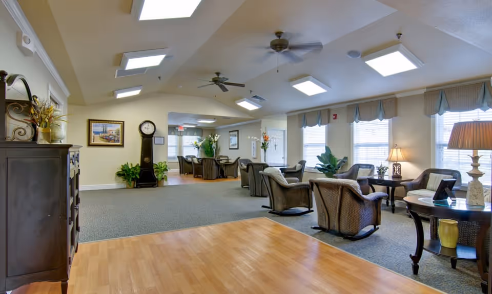 A spacious and well-lit common area in a senior living facility featuring multiple seating arrangements with wicker chairs and small tables. The room has large windows with valances, ceiling fans, and recessed lighting. There is a wooden cabinet with decorative items on the left, a grandfather clock, framed artwork on the walls, and plants placed around the room.