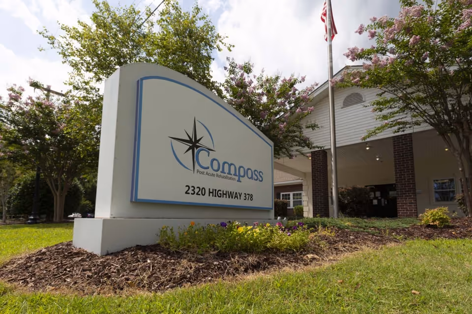 Entrance sign reading 'Compass Post Acute Rehabilitation' with the facility's front entrance, flagpole, and landscaped grounds.
