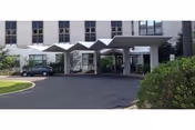 Entrance area of a senior living facility with a covered drop-off zone featuring a zigzag roof design. There are cars parked near the entrance, and the building has multiple windows. Shrubs and greenery are visible around the driveway.