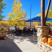Outdoor patio area with several people sitting under blue and beige umbrellas near a body of water. There are trees with yellow leaves in the background and pumpkins placed on a stone ledge in the foreground.