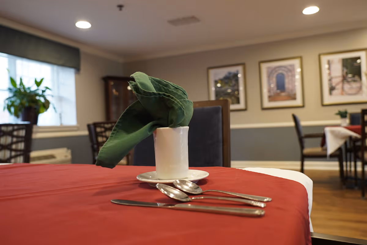A dining table set with a red tablecloth, a white cup holding a green folded napkin, and silverware including a knife, fork, and spoon. In the background, there are chairs, framed pictures on the wall, a window with a plant on the windowsill, and soft lighting from ceiling fixtures.