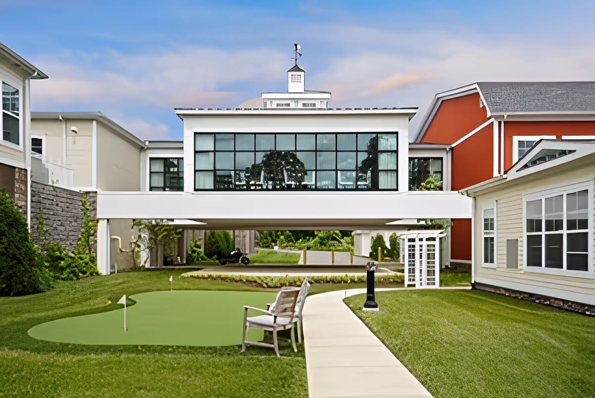 Outdoor view of a senior living facility courtyard with a small putting green, a wooden bench, a paved walkway, and well-maintained grass. The buildings surrounding the courtyard have large windows, with one building featuring a red exterior and another with a white exterior. The sky is partly cloudy.