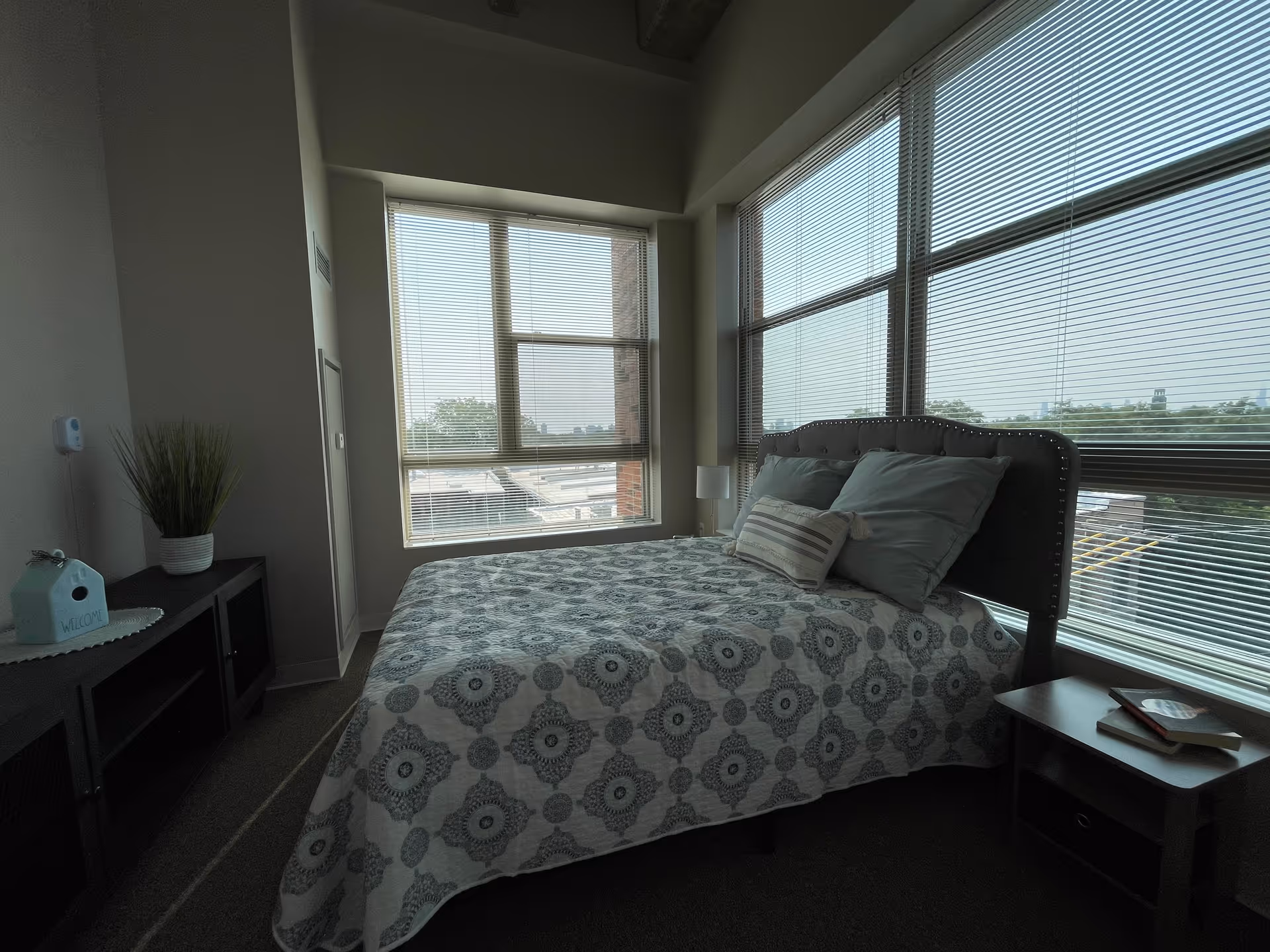 A bedroom with a bed covered in a patterned quilt and pillows, positioned near large windows with blinds. There is a small nightstand with books on the right side of the bed and a cabinet with a plant and a decorative birdhouse on the left side. The room has carpeted flooring and light-colored walls.