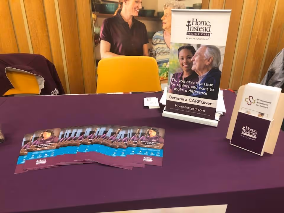 A Home Instead Senior Care information table with brochures, a tabletop banner and pamphlet holder on a purple tablecloth.