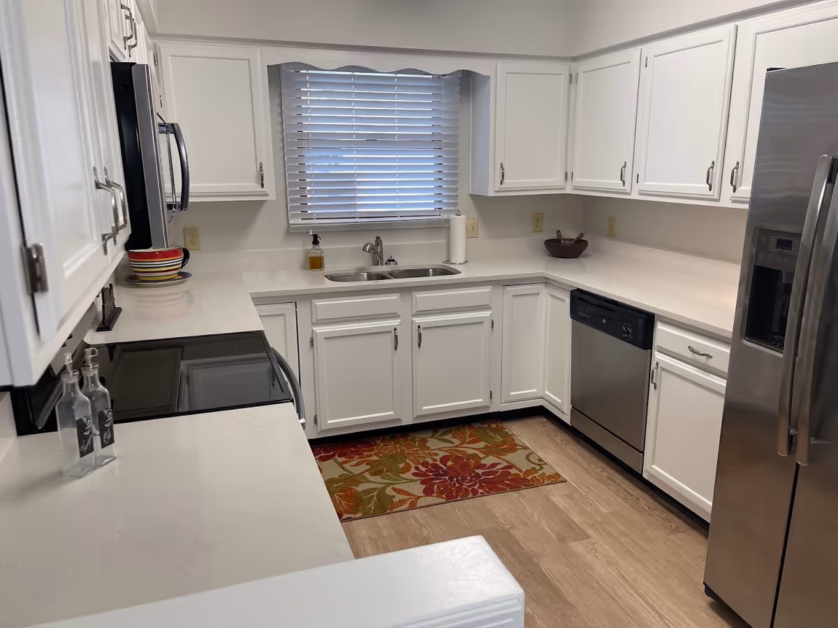 A modern kitchen with white cabinets and countertops, stainless steel appliances including a refrigerator, dishwasher, microwave, and stove. There is a window with white blinds above the sink, a colorful floral rug on the wooden floor, and various kitchen items such as bowls and soap dispenser on the counters.
