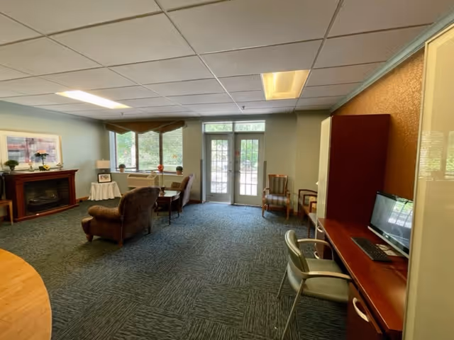A senior living facility common room with upholstered chairs, a fireplace, a computer workstation, and glass doors leading outside.