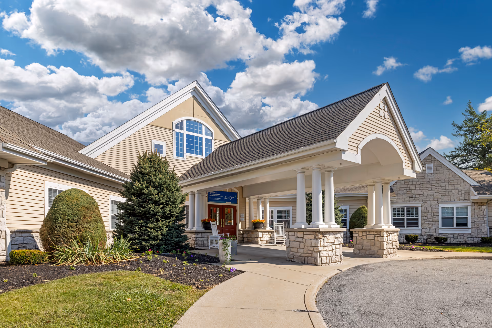 Exterior view of Brookdale Williamsville senior living facility entrance with a covered driveway supported by white columns, beige siding, stone accents, and a clear blue sky with scattered clouds.