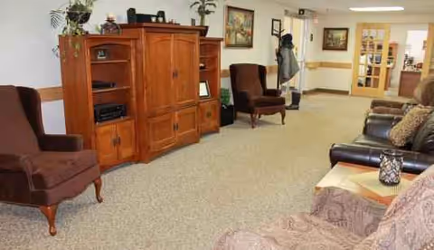 Cozy senior living lounge with armchairs, sofa, wooden cabinet, and a resident in background.