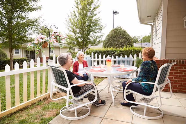 Three elderly women sitting around a round outdoor table on a patio, enjoying a sunny day. The table has a pitcher of lemonade and three glasses. The patio is surrounded by a white picket fence, with green grass, trees, and bushes in the background.